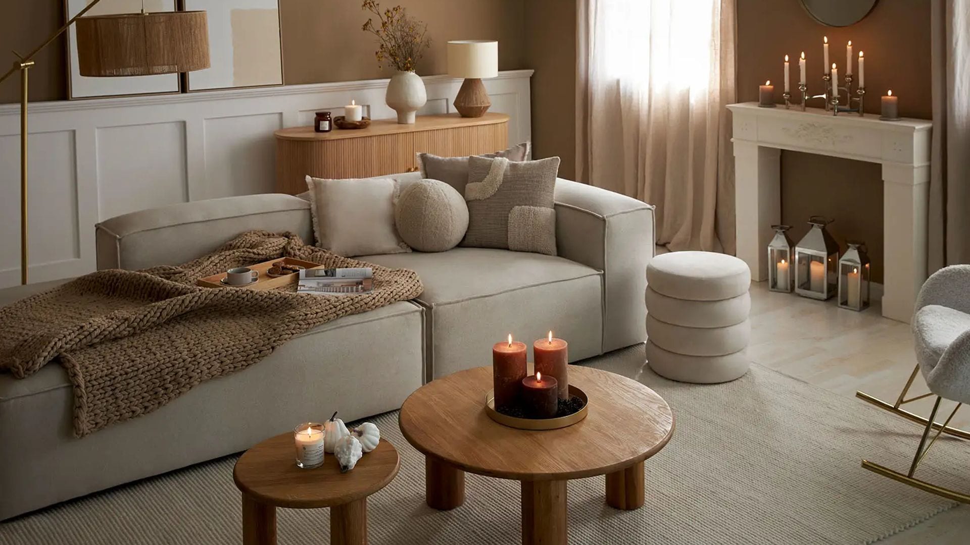 Salón acogedor en beige, madera con muchas velas repartidas por las dos mesas auxiliares, la chimenea decorativa y las repisas. Hay un sofá en en centro, un puff, una consola detrás y una butaca, además de una lámpara de pie. Paredes marrón caramelo y cortinas beige hasta el suelo.