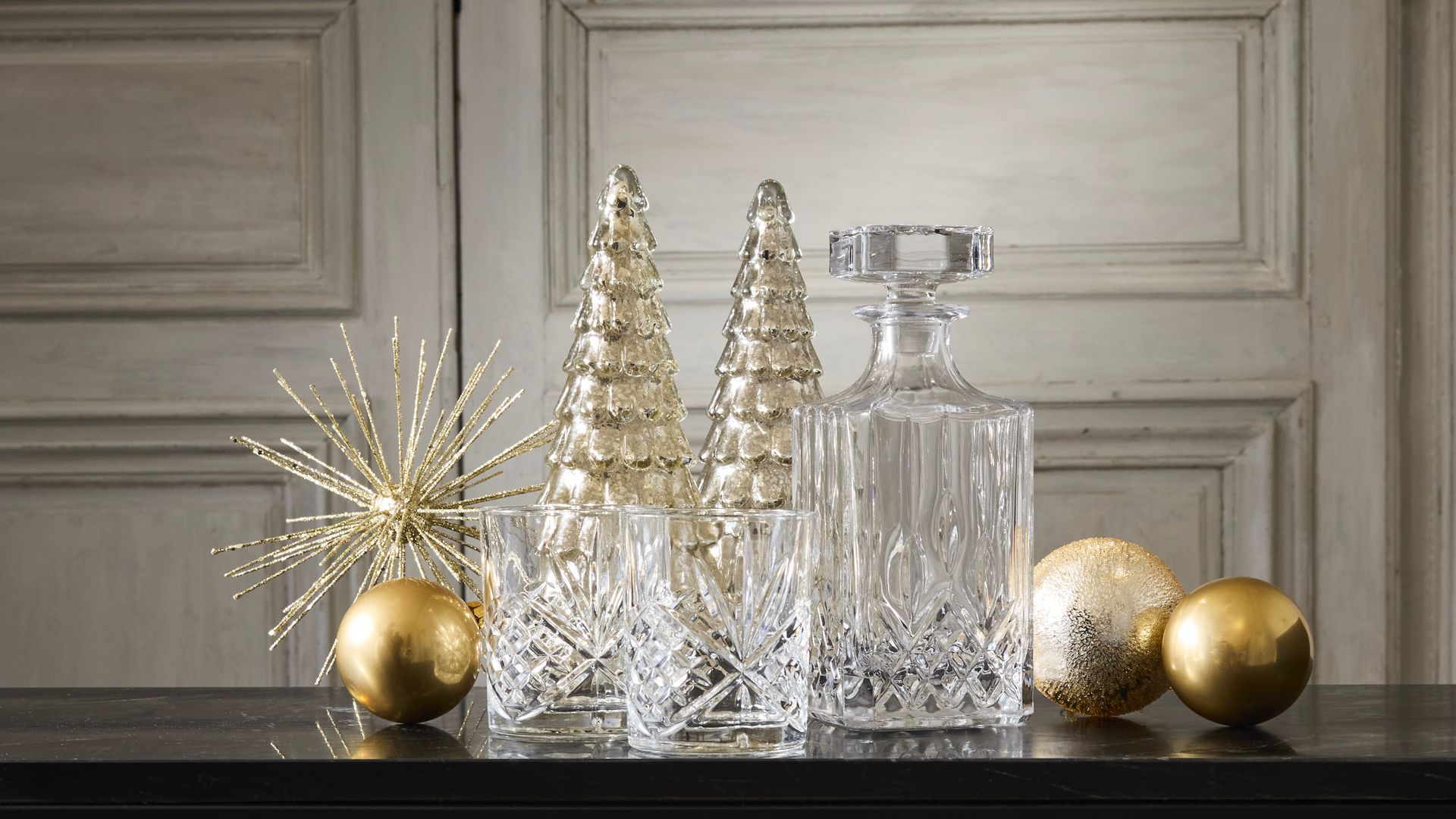 Jarra y vasos de cristal con elementos dorados de Navidad sobre una mesa negra. Maisons du Monde.