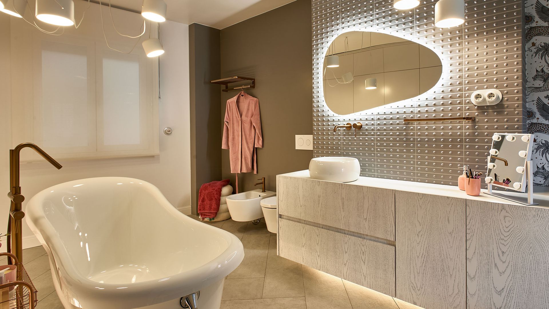 Baño con espejo de formas orgánicas retroiluminado, bañera exenta, mueble gris, lámparas de techo 