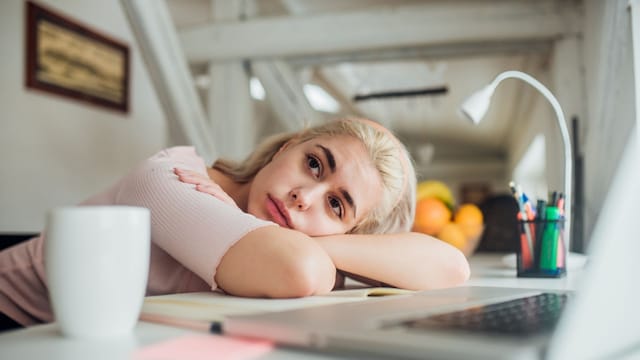 Mujer joven cansada descansando sobre el escritorio, con una taza de café blanca sobre la mesa y un ordenador portátil. Ella es rubia y va vestida de rosa.
