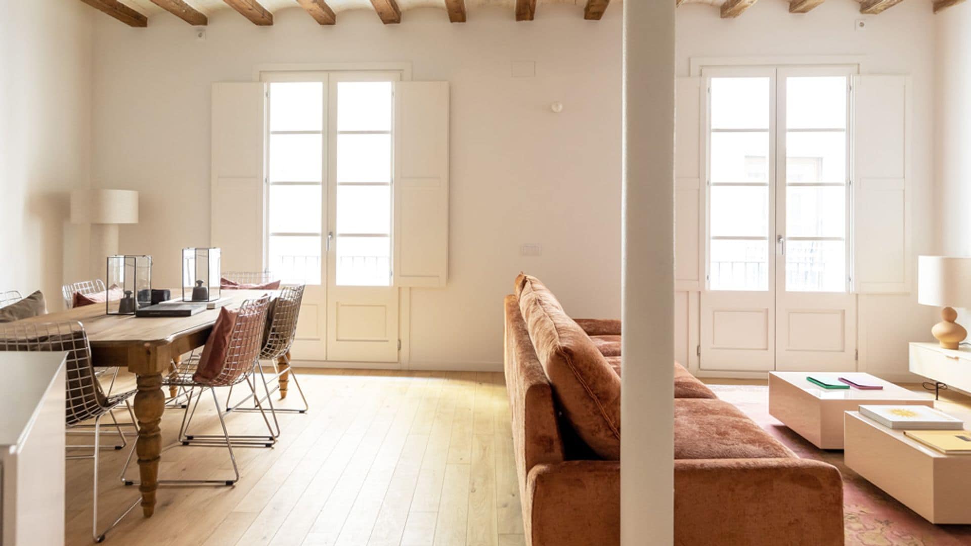 Salón comedor con vigas de madera, ventanas mallorquinas blancas, suelo de madera, sofá marrón, mesa de madera