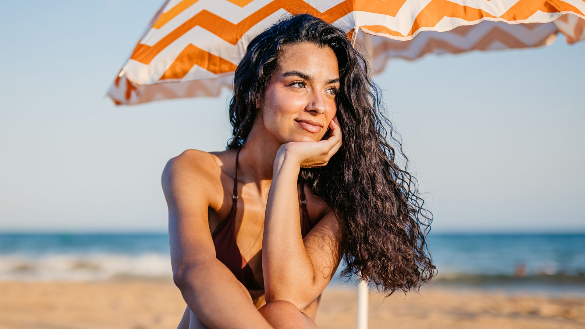 Mujer joven morena sentada en la playa en bikini con sombrilla estampada y melena rizada suelta