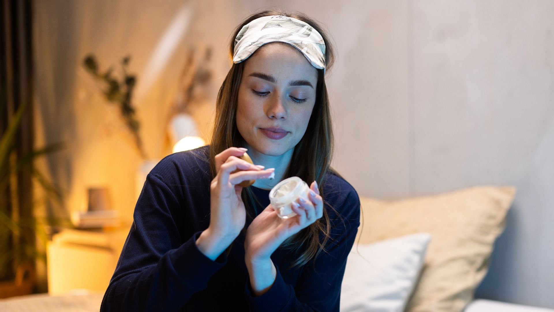 Mujer en pijama sentada en la cama con antifaz aplicándose crema facial