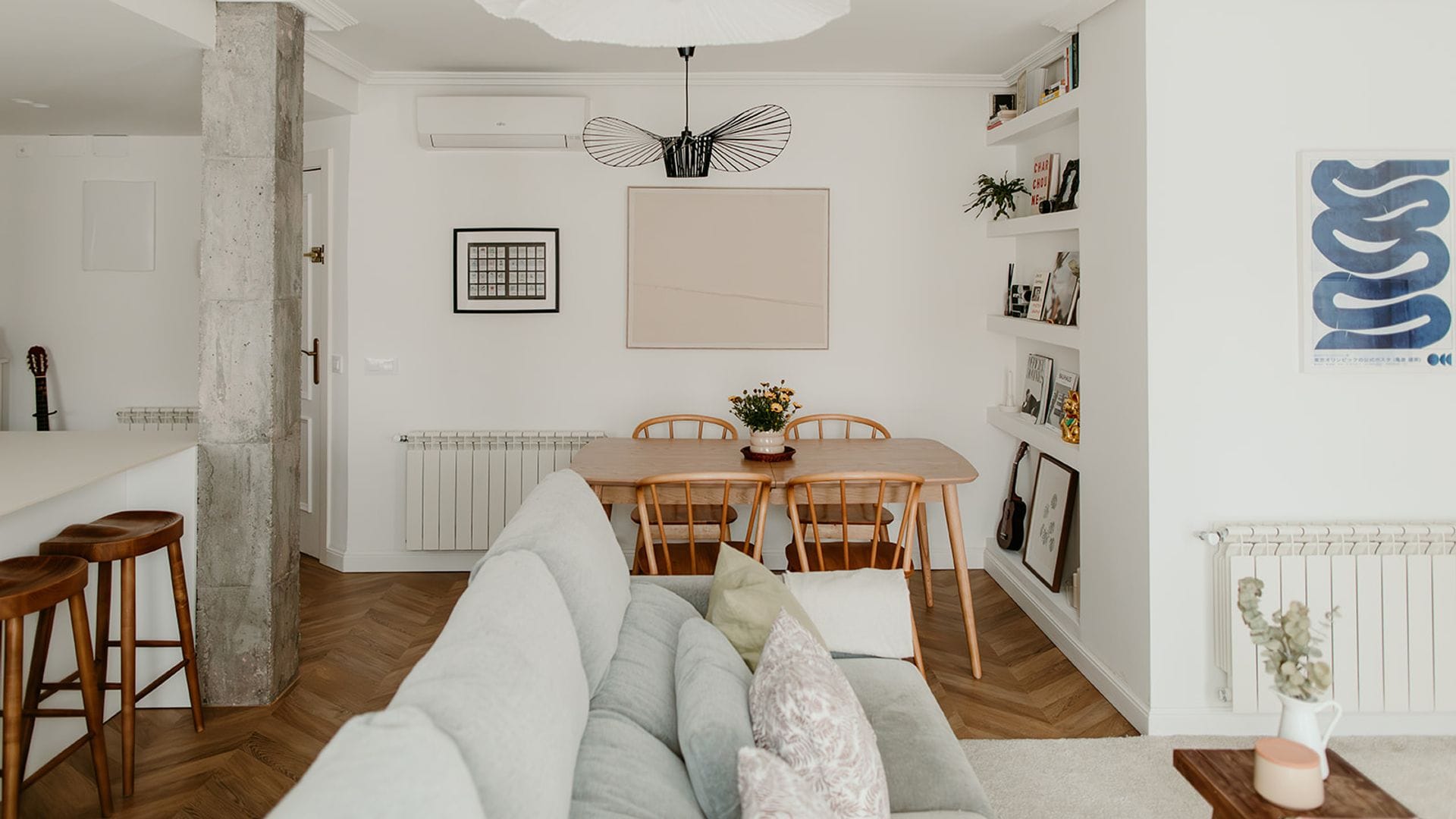 Salón comedor y cocina en un mismo espacio, con suelo de madera en espiga, mesa de madera con sillas, columna de cemento, taburetes altos, cuadros, lámpara de techo blanca, radiadores blancos,, librería de obra