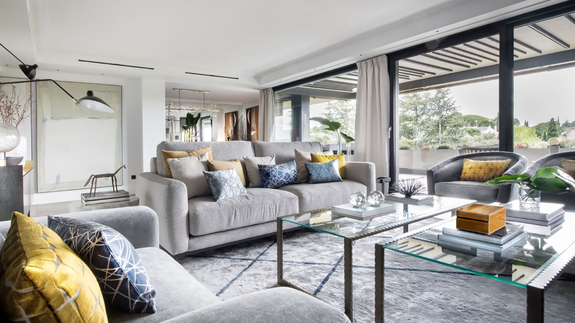 Salón con sofás grises, alfombra gris, ventanales con salida al jardín, cortinas blancas, mesas de centro de cristal, cojines de colores en los sofás 