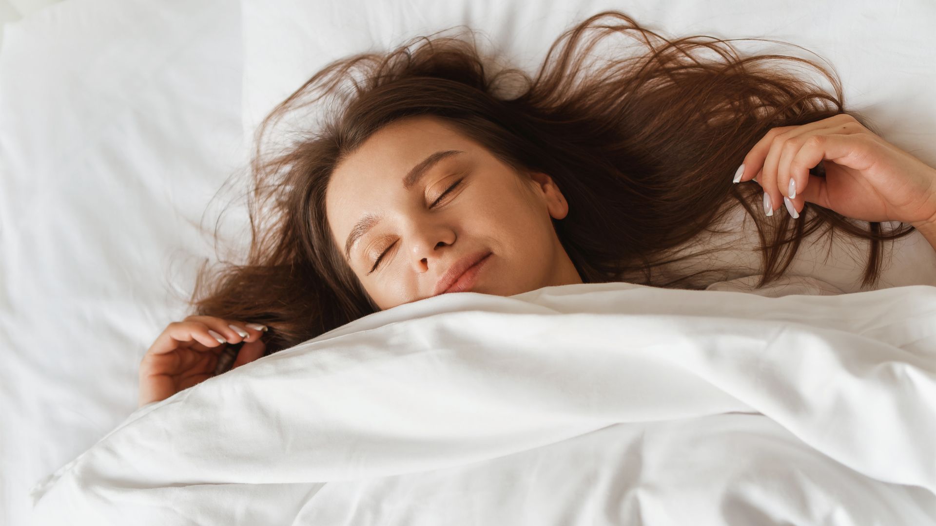 Mujer joven durmiendo en una cama de sábanas blancas con el pelo suelto, boca arriba y sonriente.