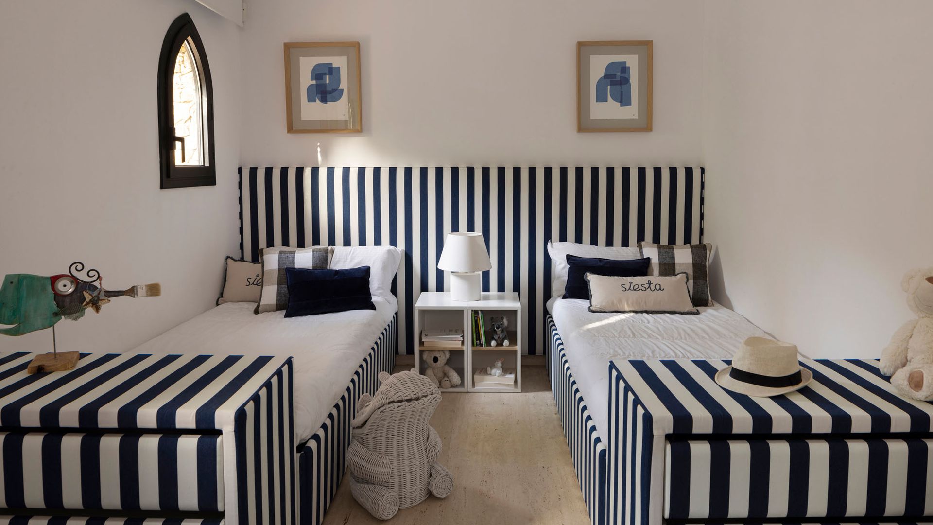 Habitación infantil con dos camas de rayas negras y blancas, cabecero a juego, cuadros en la pared, mesita de noche blanca, lámpara de sobremesa
