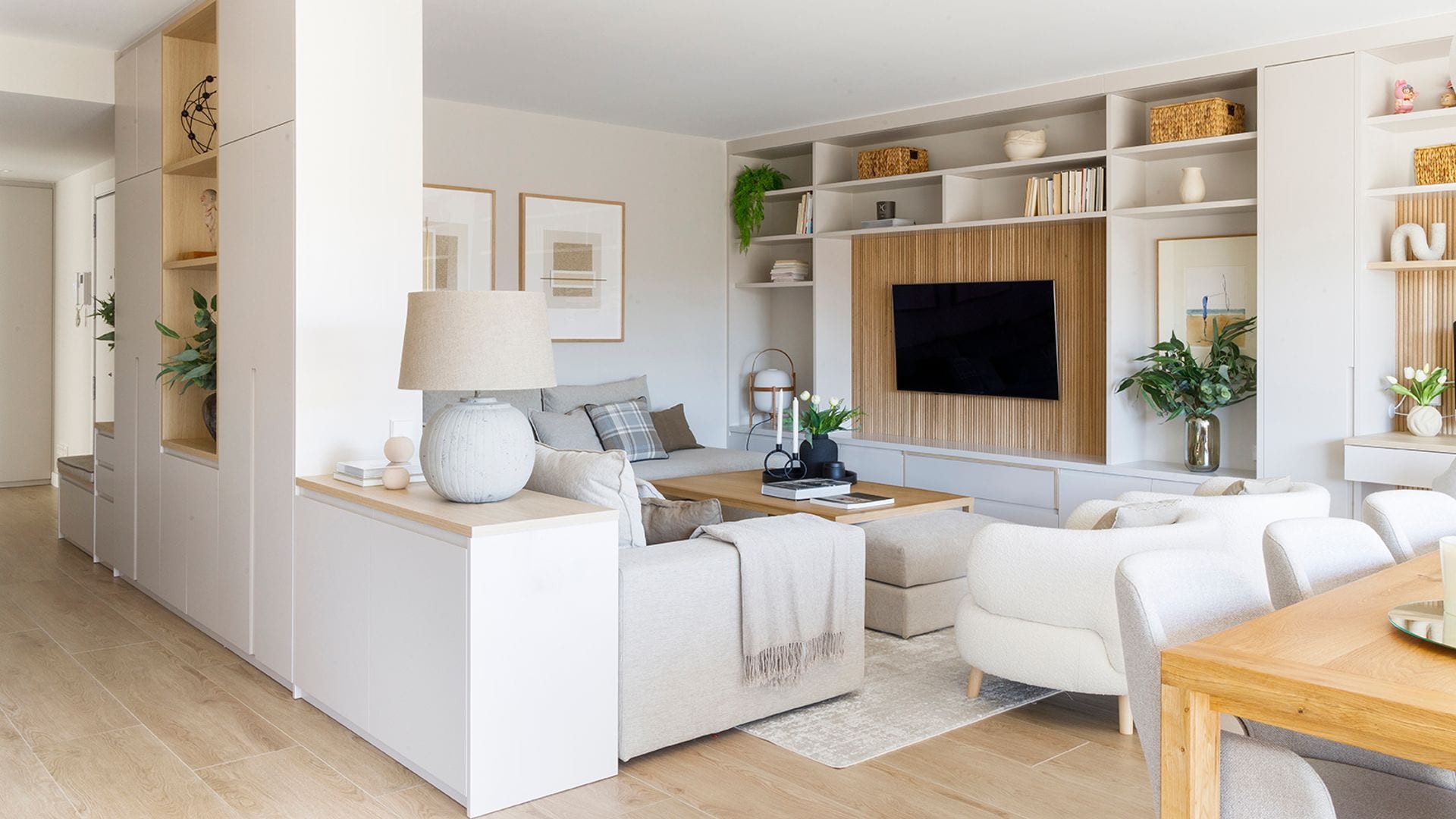 Salón comedor abierto al recibidor, con mueble a medida hasta el techo blanco con una parte abierta, estantería a medida hasta el techo con la televisión colgada, sofás beiges