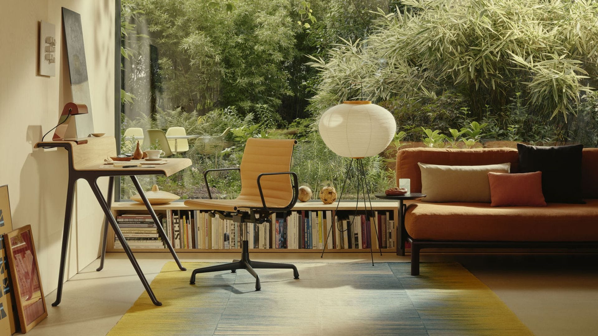 Salón con zona de despacho, estantería baja, ventanal con vistas al jardín, alfombra, sofá marrón con cojines