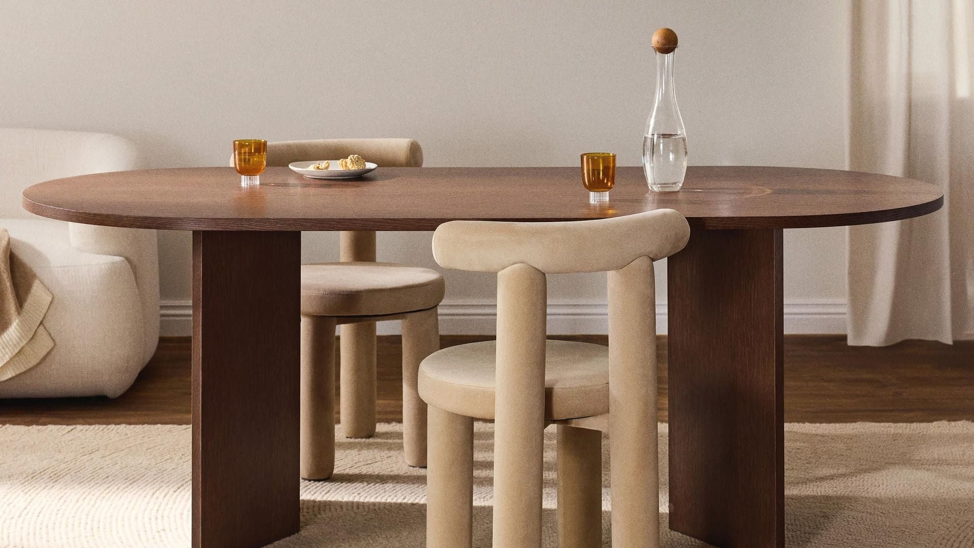 Comedor en tonos tierra, con mesa de madera ovalada, dos sillas y una lámpara baja en madera. Materiales naturales. Otoño-invierno 2025. Westwing.