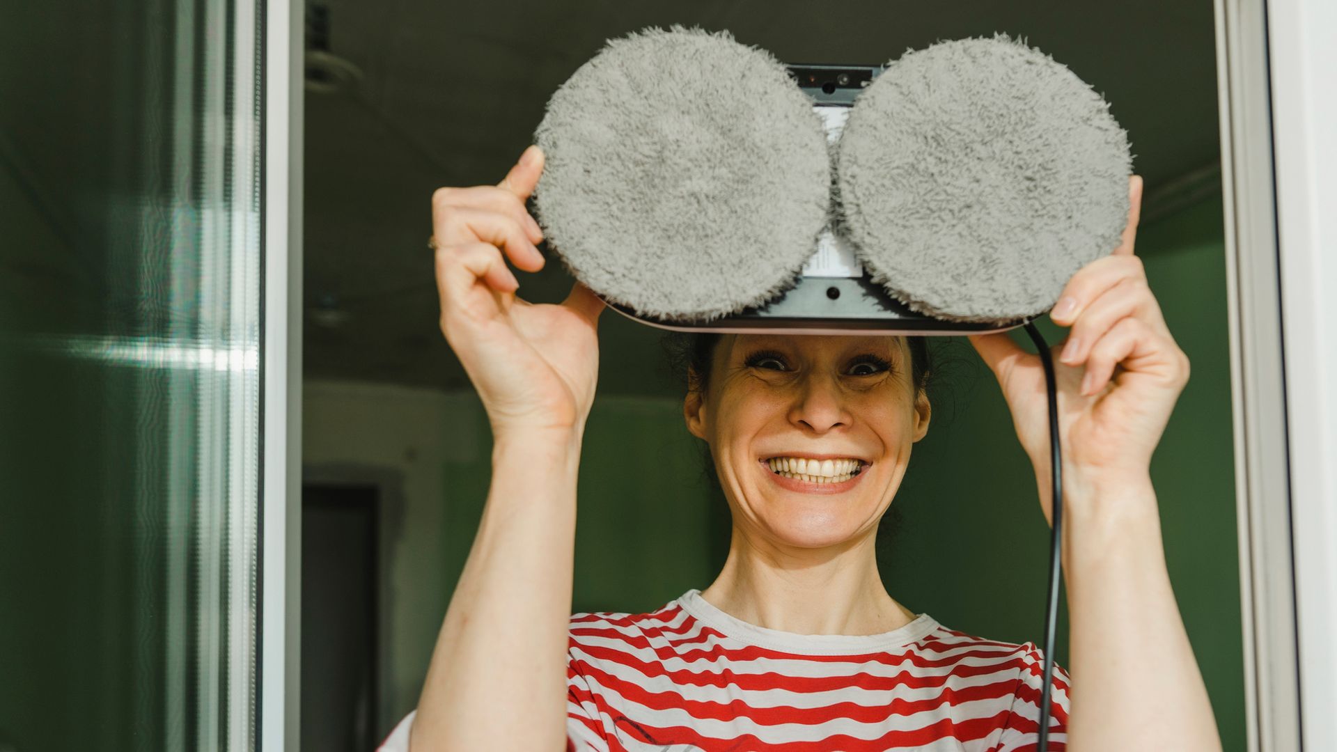 Mujer feliz con un robot limpiacristales en las manos al lado de una ventana y mirando de frente.