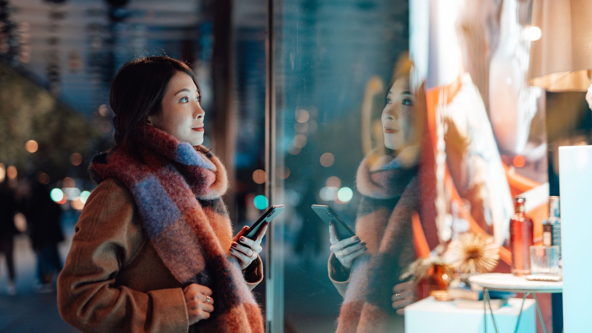 Mujer joven comprando en un centro comercial, mirando un escaparate desde fuera, con ropa de invierno y teléfono móvil en la mano.