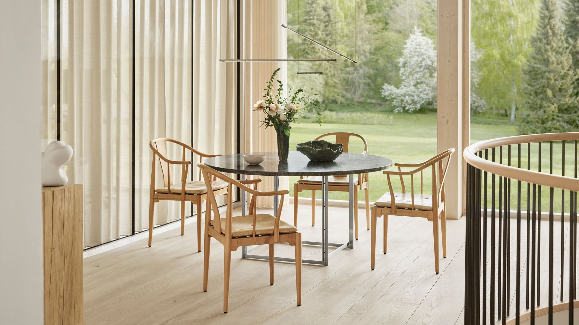 Comedor con mesa redonda, sillas de madera, grandes ventanales con salida al jardín, jarrón on flores, adornos negros y blancos sobre la mesa