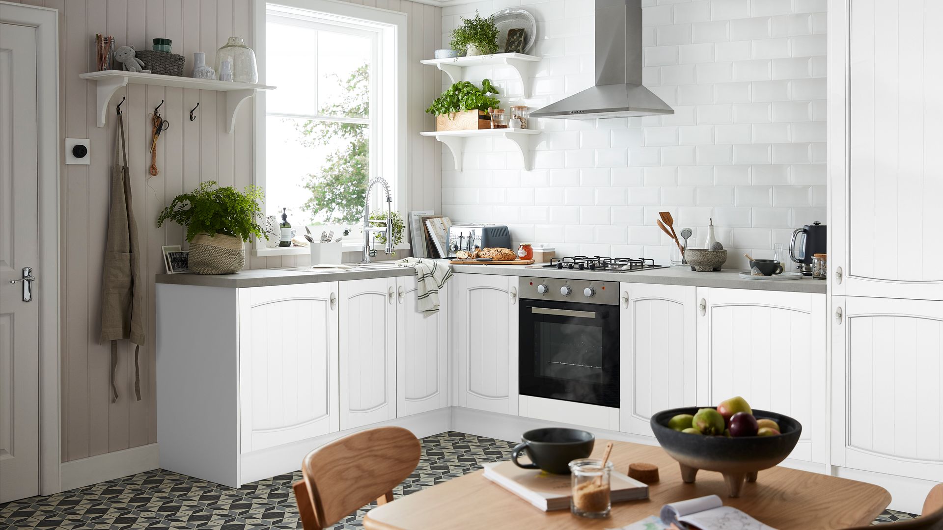 Cocina blanca con pared de azulejos tipo metro, estanterías con plantas, perchero con estantería, suelo de baldosas, mesa de comedor con frutero y taza, 