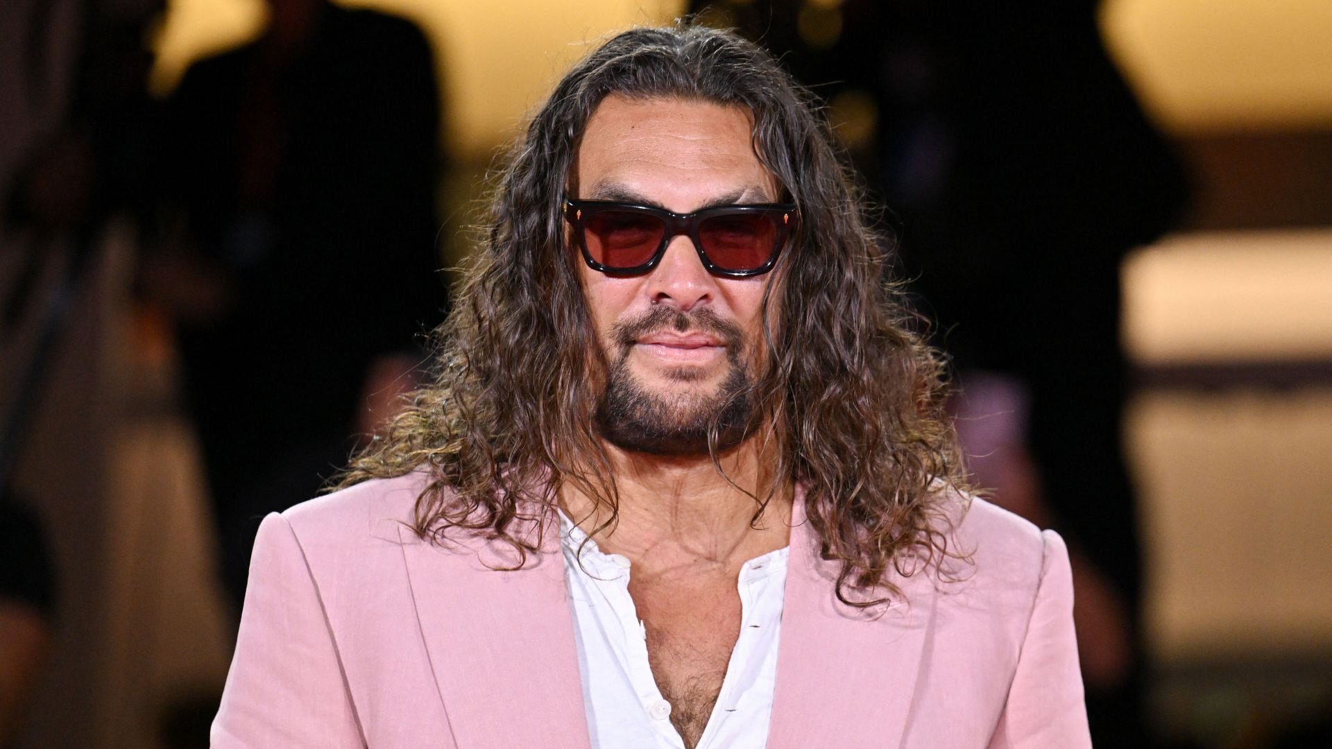 Jason Momoa con cabello largo, barba, gafas de sol y traje rosa.