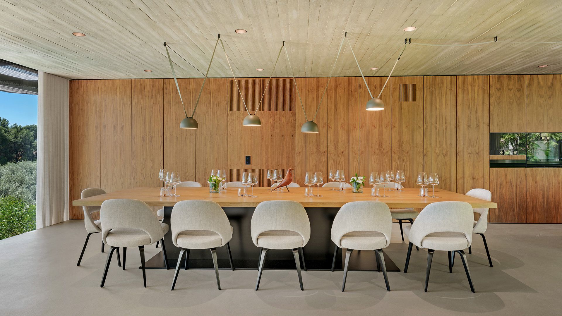 Comedor con mesa XL de madera, sillas tapizadas en beige, lámparas de techo sobre la mesa, pared de armarios de madera