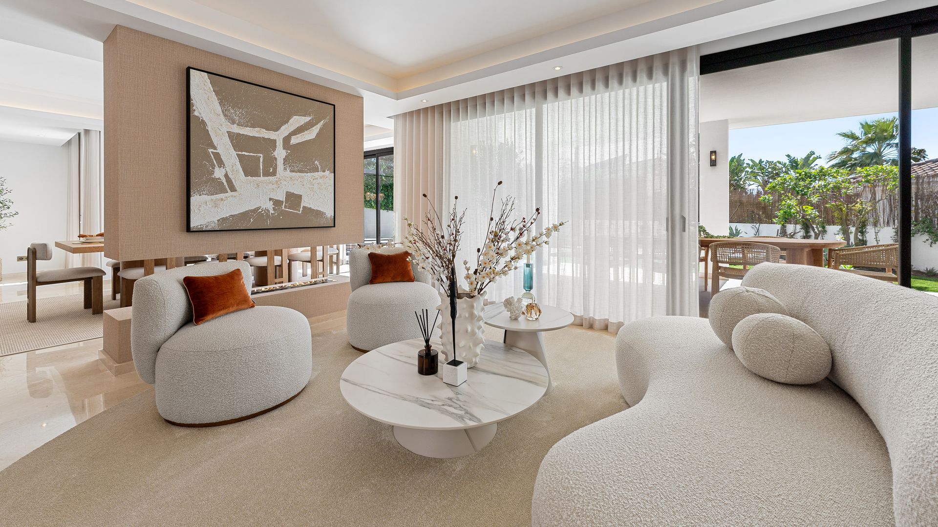 Salón en tonos beiges con sofá blanco de forma curva, alfombra, cuadro en la pared, chimenea dividiendo el espacio con el comedor, butacas blancas, mesas de centro con el sobre de mármol 