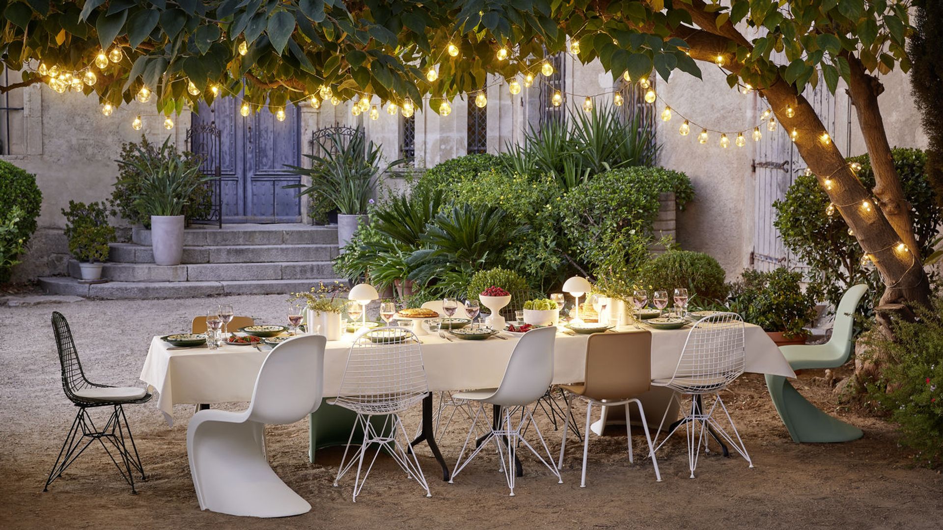 Mesa de verano bajo un árbol con guirnaldas de luz, sillas Panton, mantel blanco