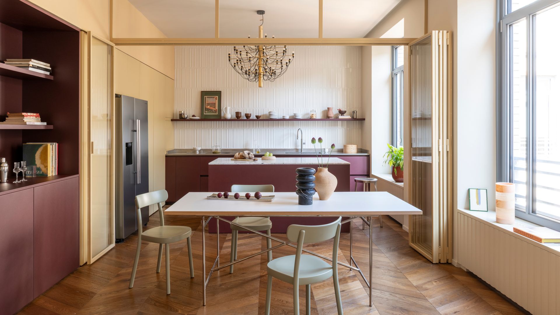 Cocina con comedor con suelo de madera, muebles de cocina de color burdeos, mesa de comedor blanca, sillas de comedor, puertas de cristal tipo acordeón entre la cocina y el comedor 