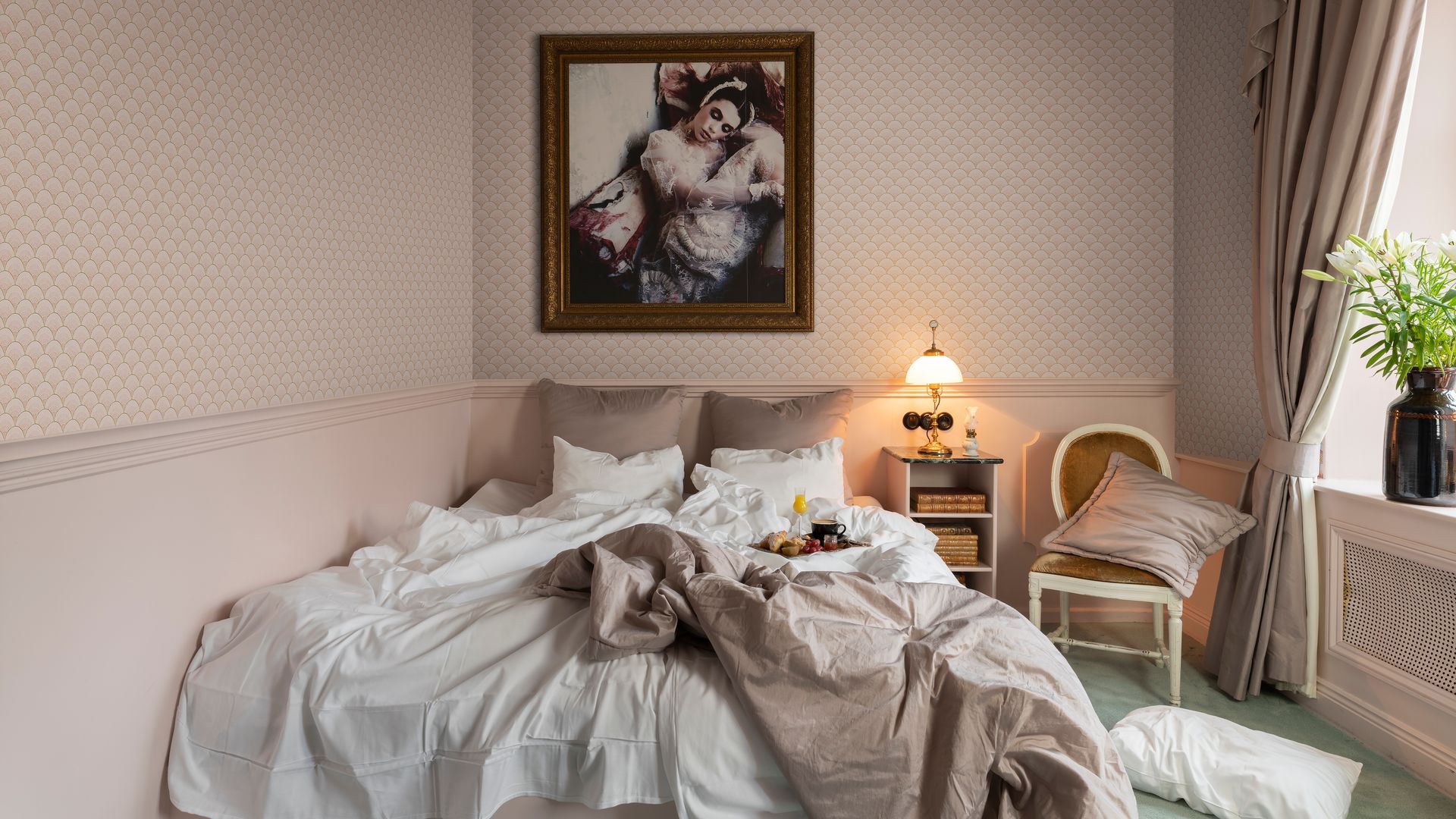 Dormitorio con papel pintado, cuadro en la pared del cabecero, zócalo alto, cama con bandeja desayuno, silla, lámpara de sobremesa sobre la mesita de noche, jarrón negro en la ventana, cubrerradiador