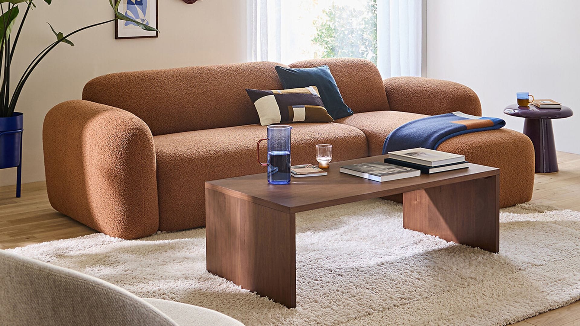 Sofá de formas curvas, lana bouclé marrón, alfombra de pelo largo beige, mesa de centro de madera, cuadros en la pared, butaca y maceta azul 