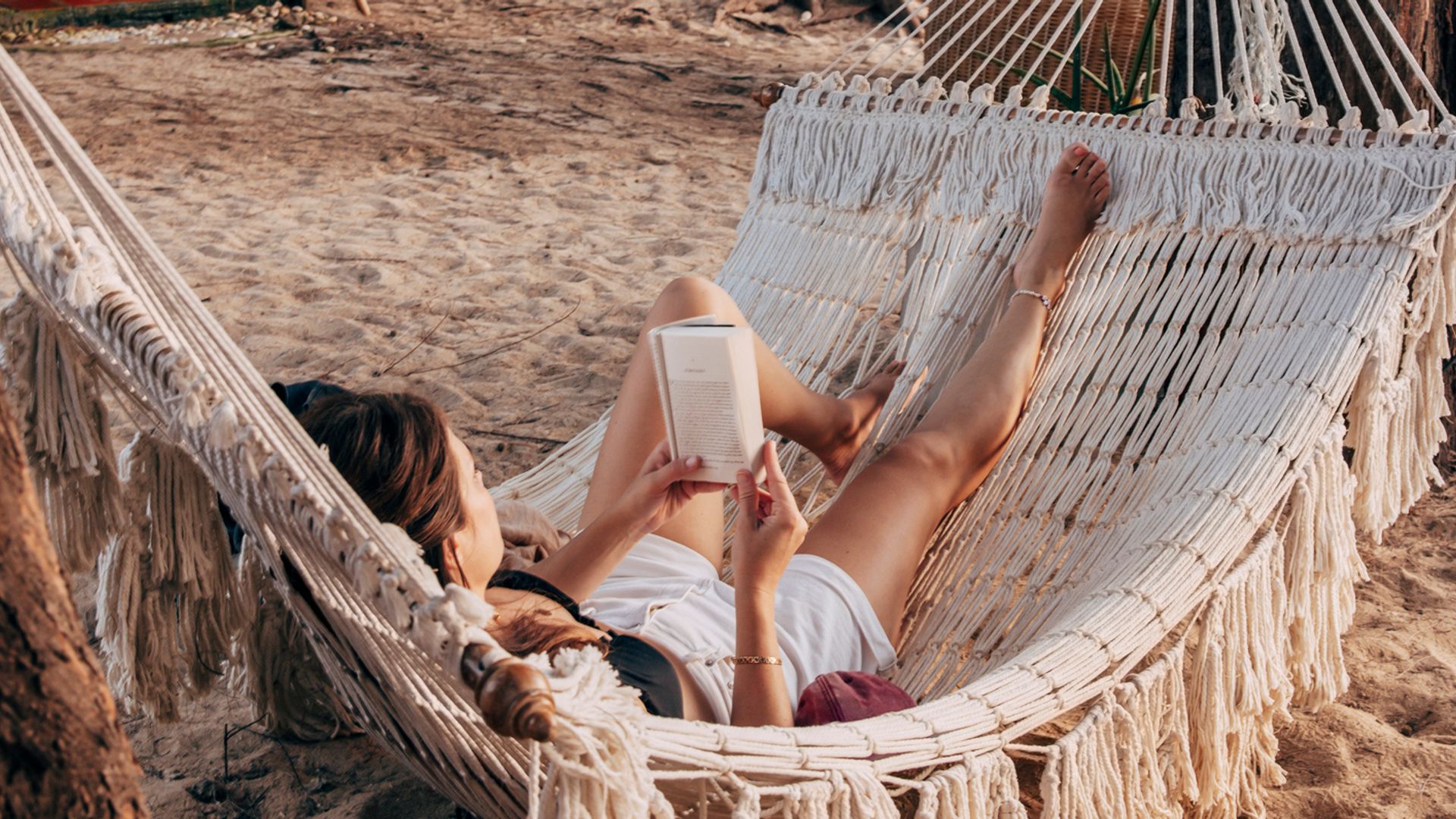 Mujer leyendo un libro tumbada en la hamaca de una playa, debajo de un pinar.