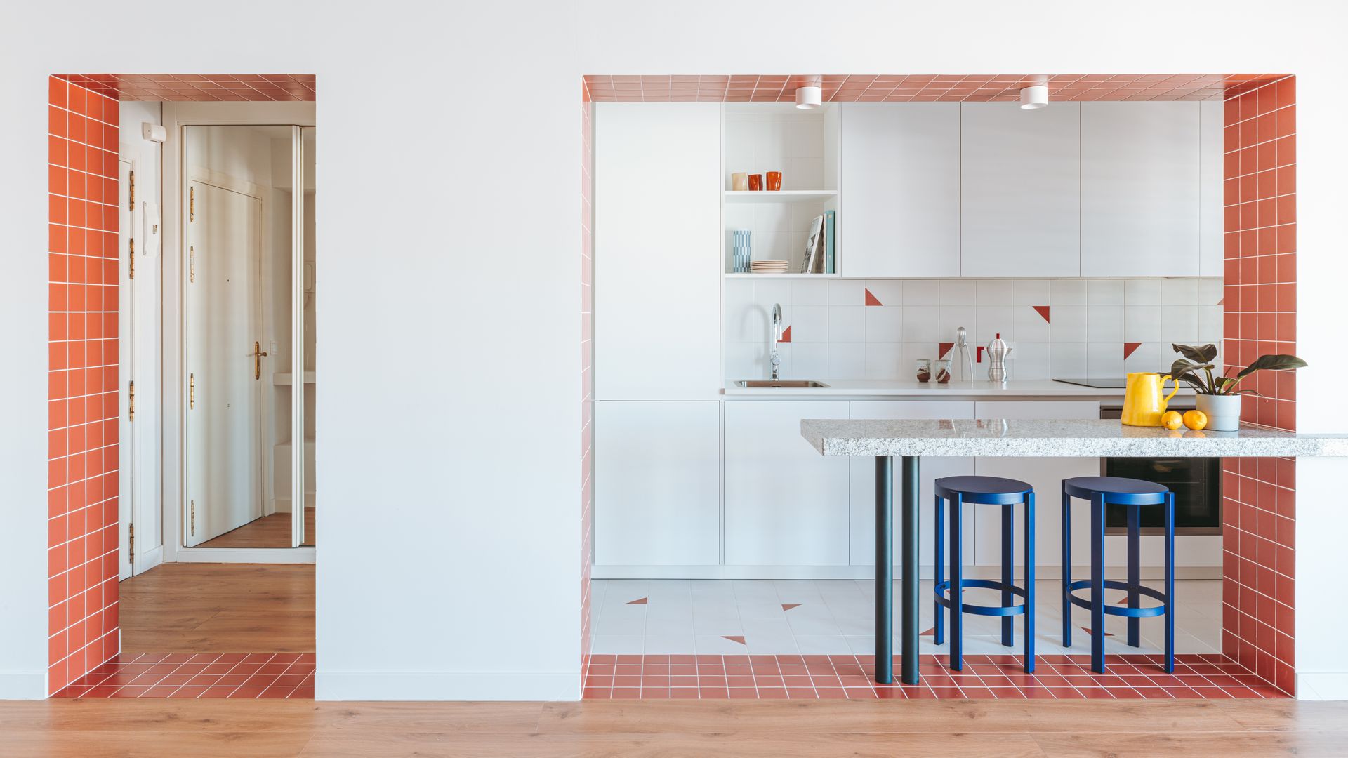 Cocina con barra, azulejos naranjas, muebles de cocina blancos, taburetes azules, suelo de madera, suelo porcelánico 