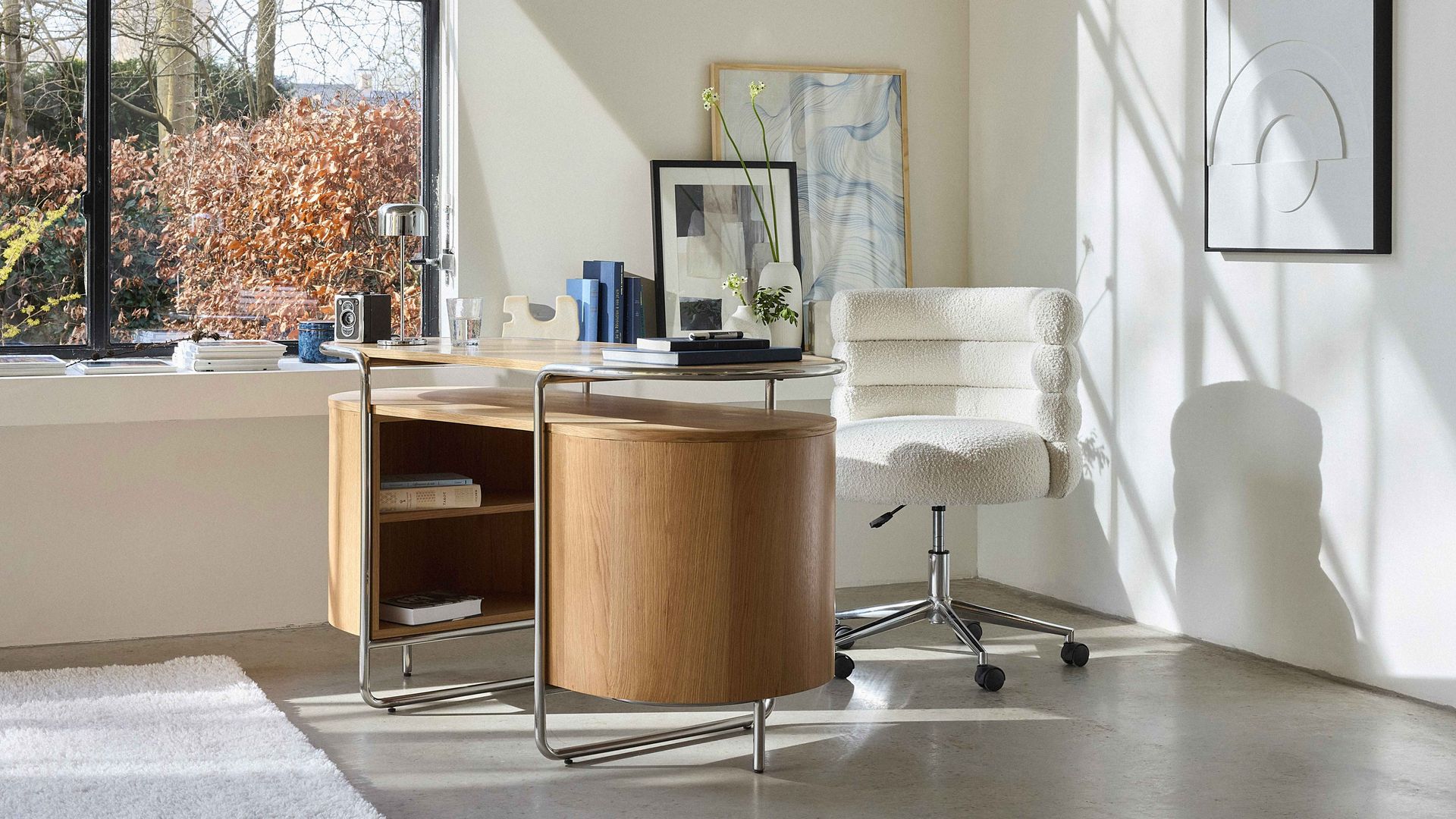 Despacho en casa con gran ventanal, mesa de madera con cajones, silla de oficina blanca con ruedas, cuadros en la pared 