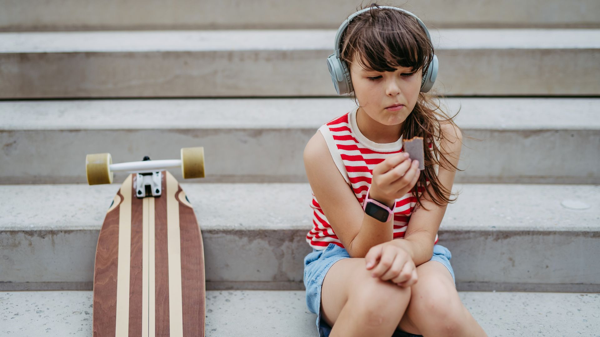 Niña con su monopatín sentada en las escaleras de la calle mientras se come una galleta, con un reloj inteligente en la muñeca y unos auriculares de diadema.
