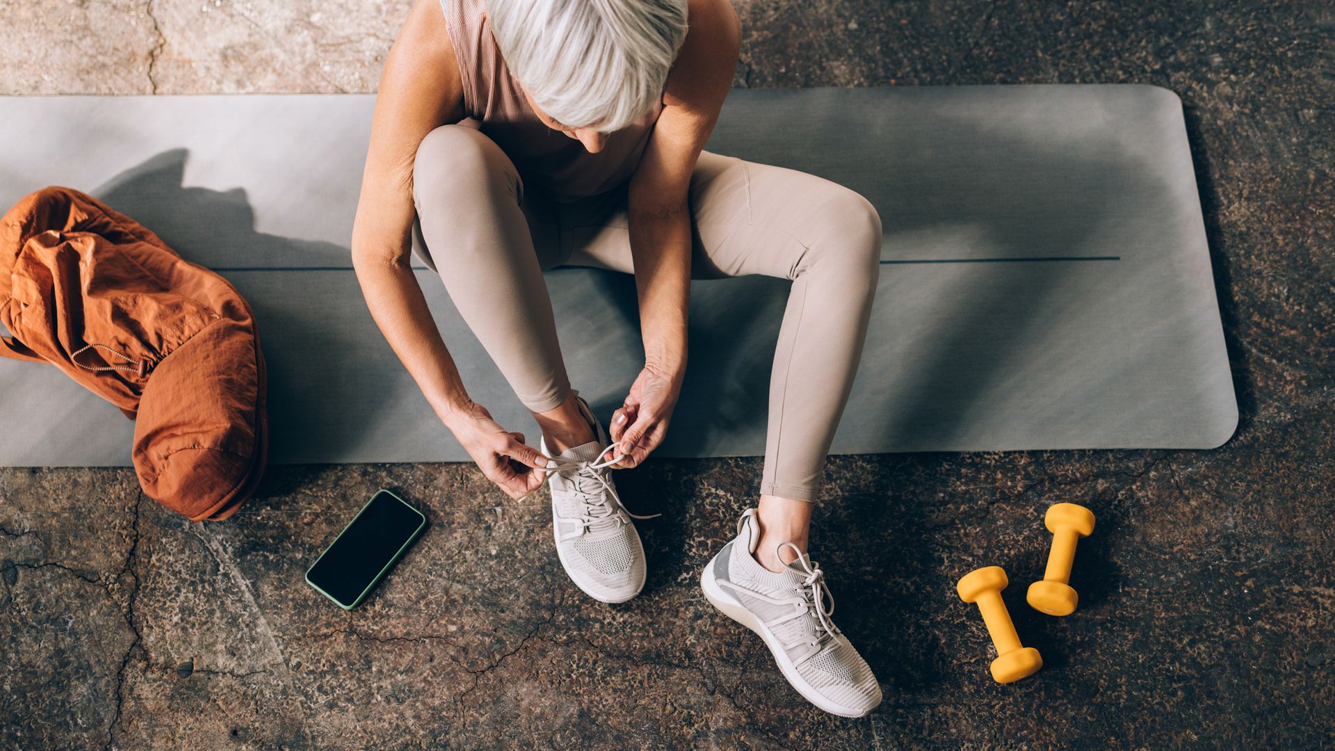 Fotografía en plano picado de una mujer de 60 años sentada en una esterilla mientras se ata las zapatillas de deporte