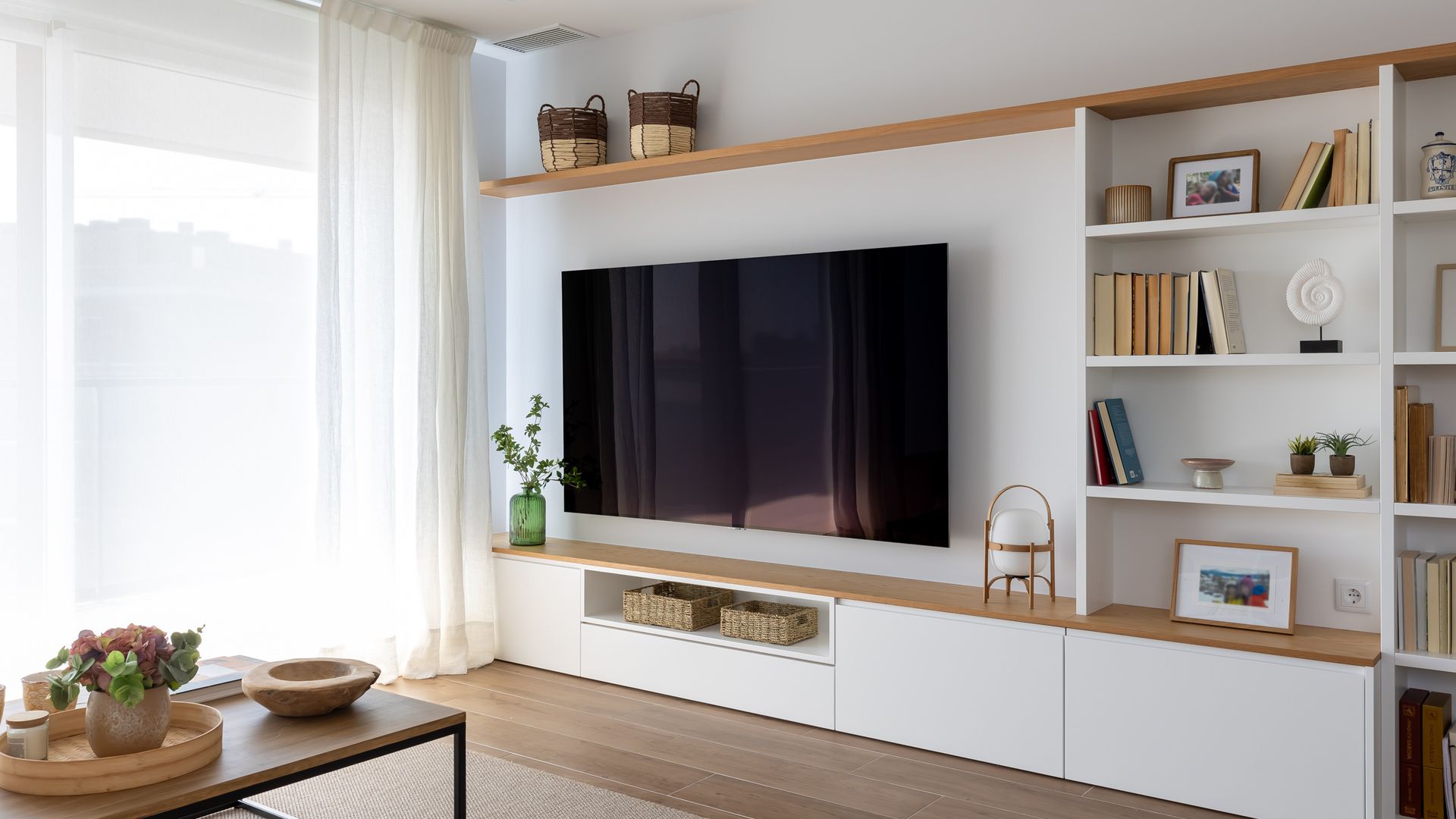 Salón con televisión colgada en la pared, mueble a media en blanco y madera con balda superior y cajones en la parte inferior, cortinas
