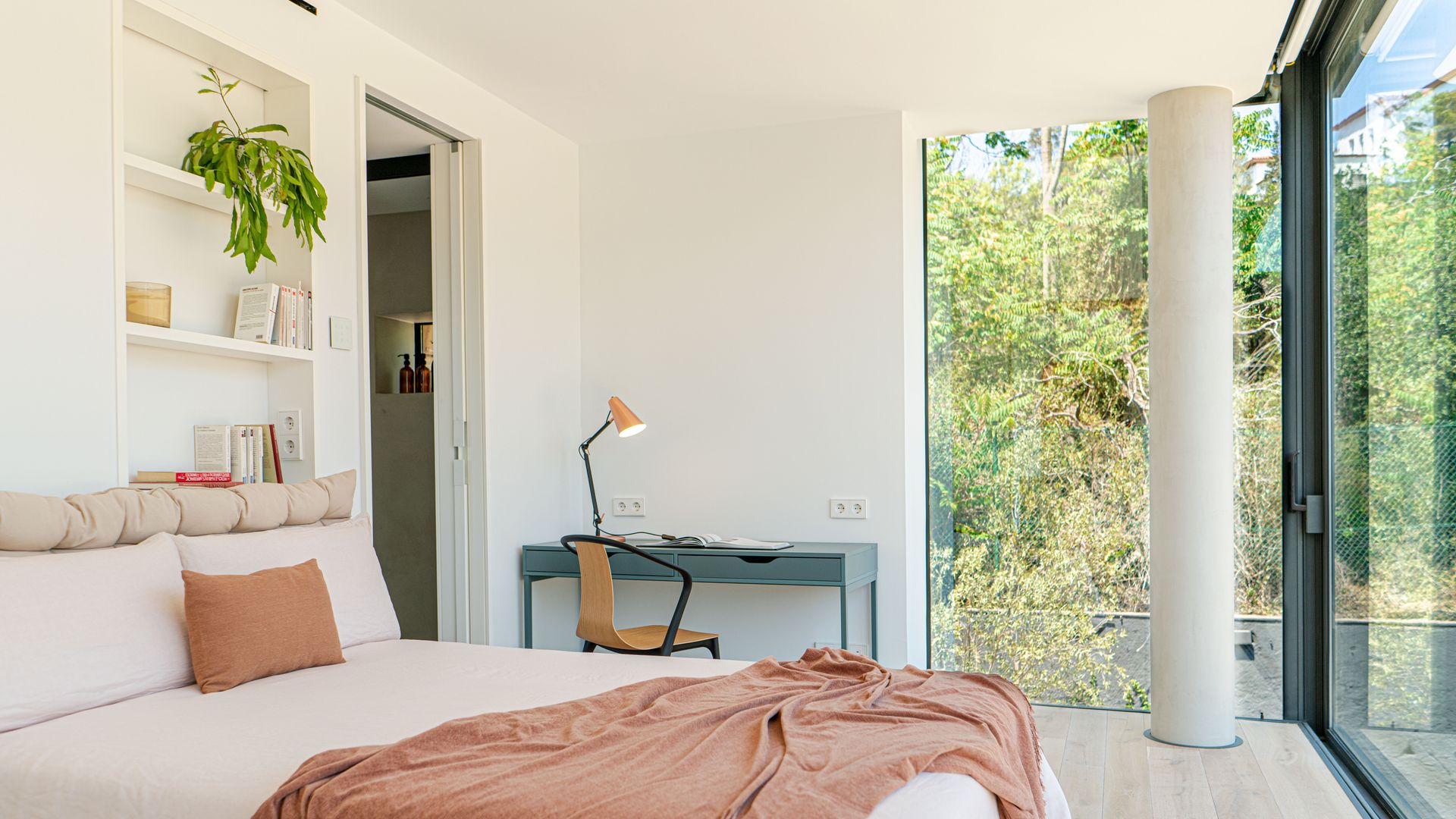 Dormitorio con cabecero de obra con estantería blanca, grandes ventanales con vistas al jardín, zona de escritorio con mesa azul grisácea y flexo, ropa de cama blanca con colcha tierra