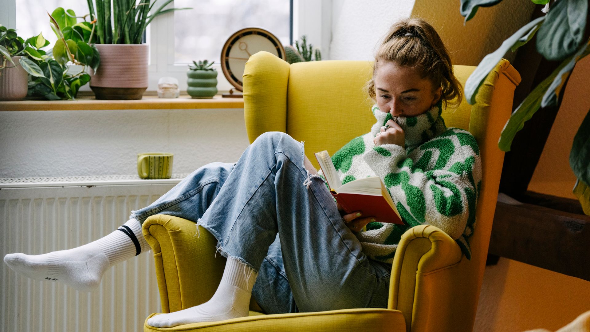 Retrato de una mujer pelirroja sentada en un sillón amarillo rodeada de plantas en su sala de estar, leyendo un libro.