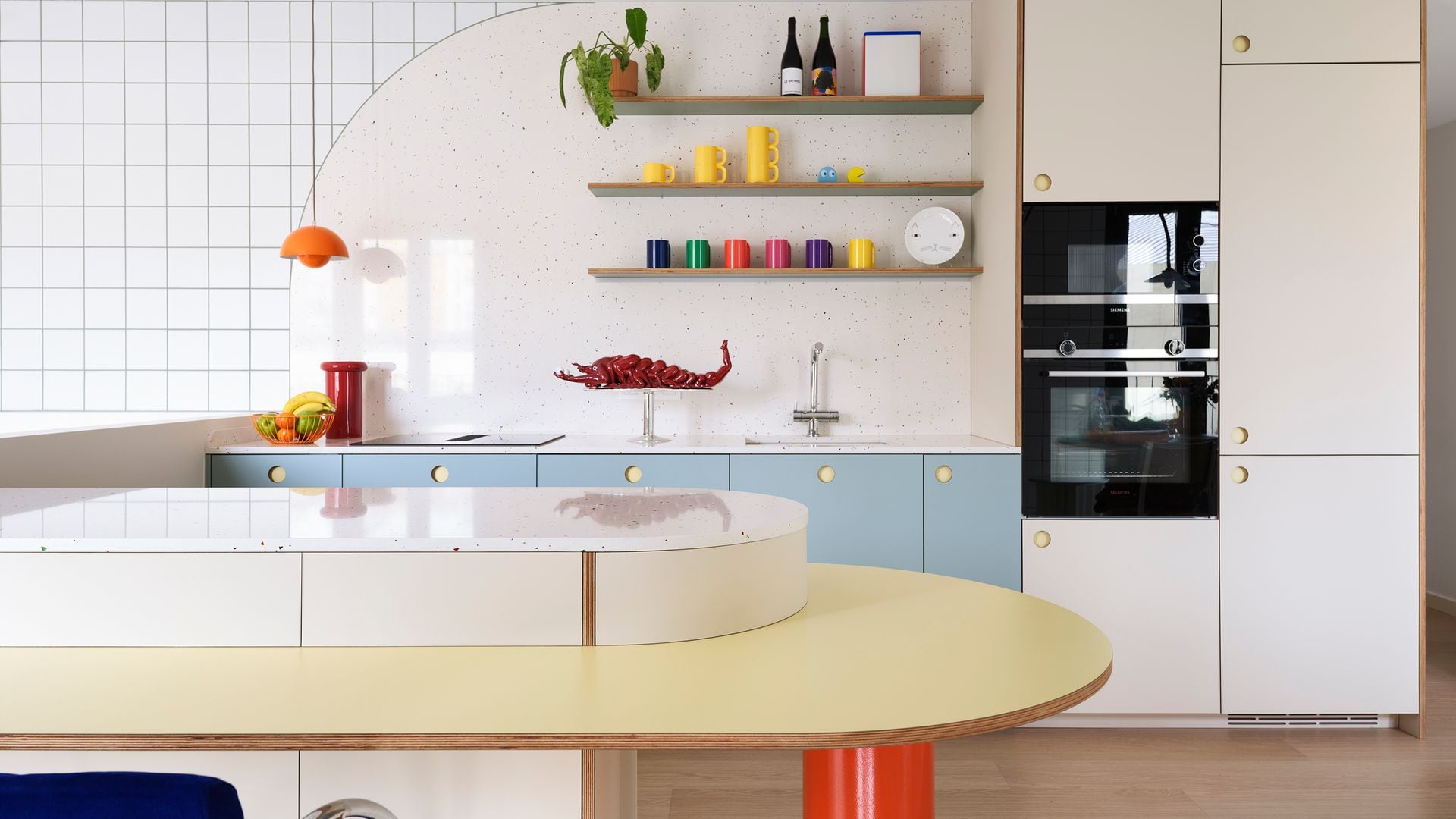 Cocina con muebles azules, barra curva amarilla, armario blanco con horno y microondas, baldas de madera con vasos y tazas de colores, lámpara de techo naranja 