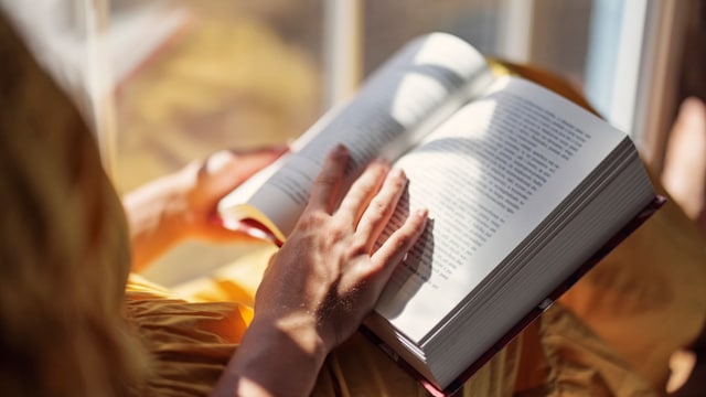 Chica leyendo un libro junto a la ventana mientras entra el sol. Tiene un libro abierto entre las manos en primer plano.