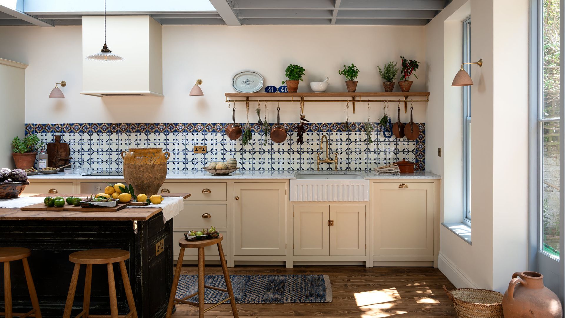Cocina de aire rústica con muebles en color beige, antepecho de azulejos azules, mesa de cocina de madera, taburetes, cazuelas y sartenes de cobre colgando de una barra