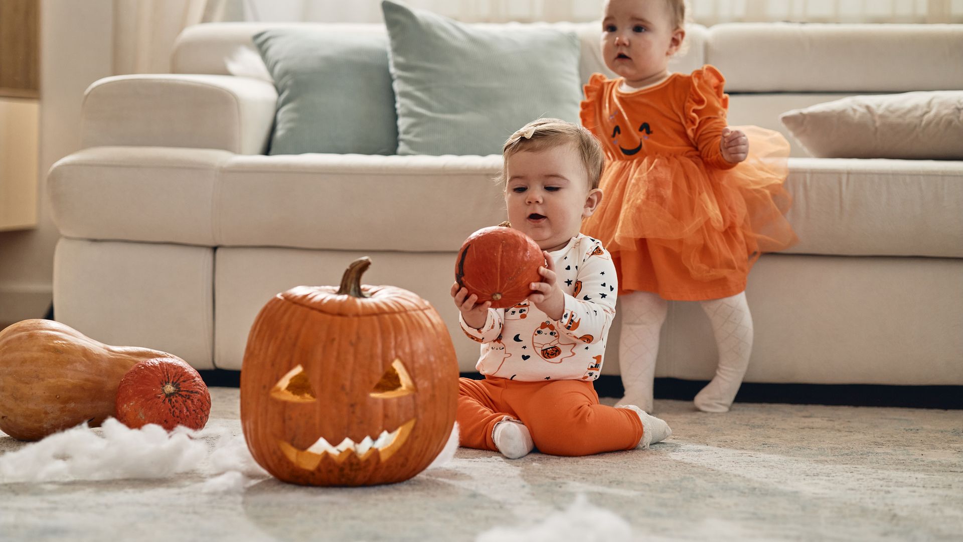 Dos bebés niñas en Halloween, en la alfombra del salón jugando de calabazas y disfrazadas también de ellas. Una está de pie junto al sofá beige del fondo y la otra sentada delante con una calabaza en las manos.