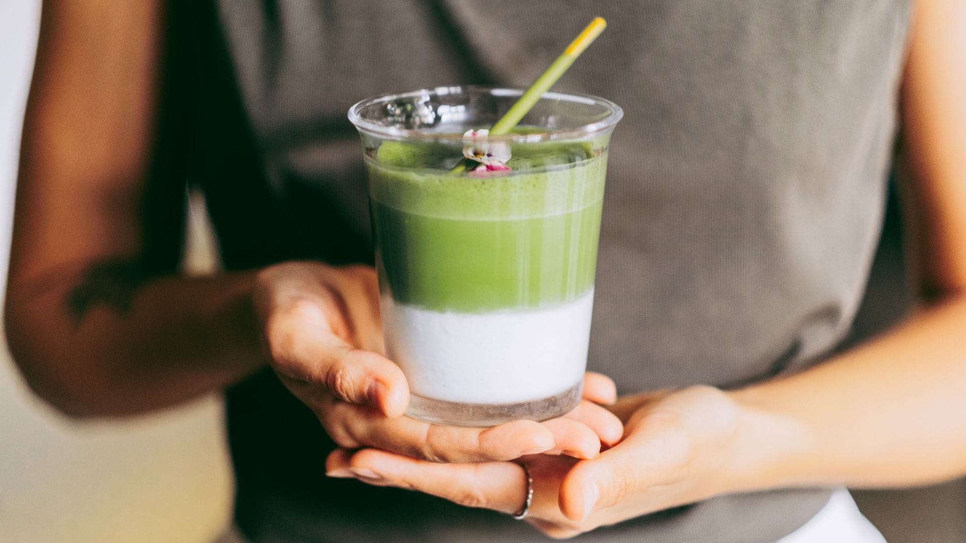 Un vaso de té matcha con leche en las manos de una chica que lleva una camiseta marrón y pantalones blancos. Es un plano detalle del vaso con una pajita.