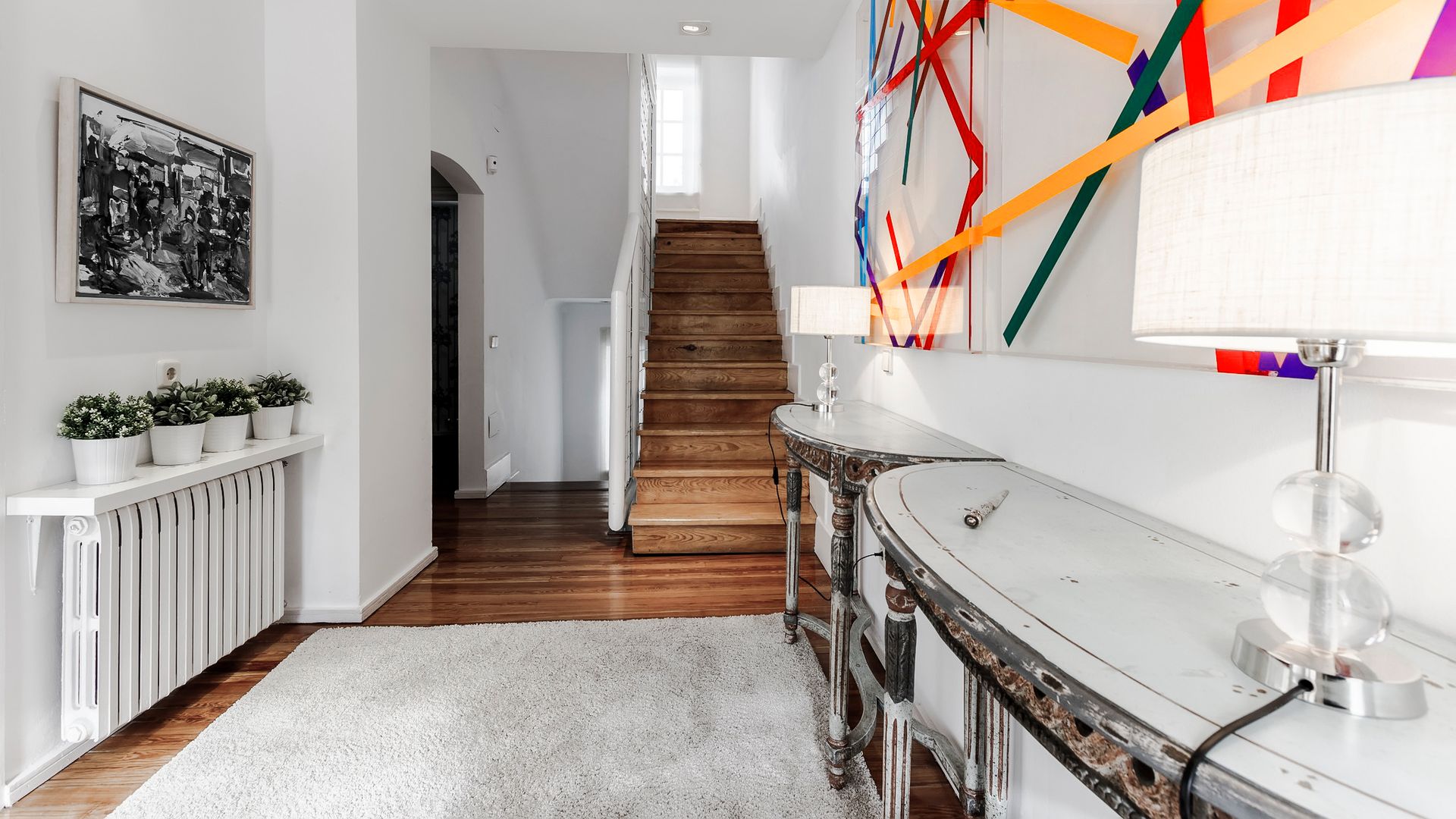 Recibidor con subida escalera de madera, dos consolas, alfombra gris clara, cuadro en la pared, radiador con repisa con macetas, lámparas de sobremesa