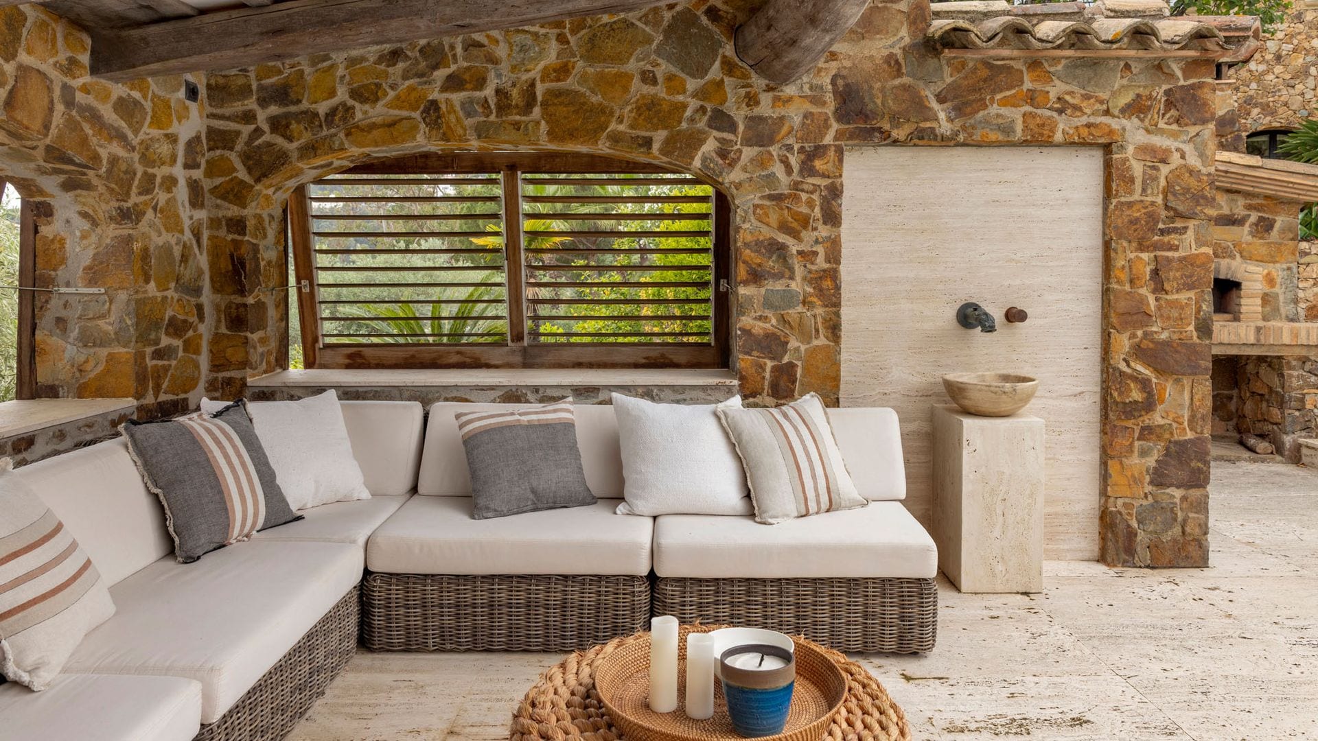 Salón exterior bajo porche de piadra, con sofás de fibras vegetales con almohadones blancos, mesa de centro, velas, fuente