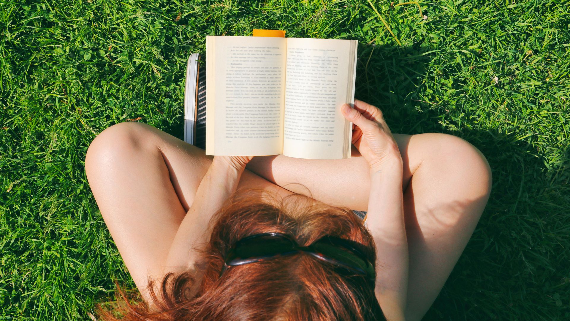 Mujer joven leyendo en el césped de un parque en pantalones cortos con gafas de sol, muy veraniega. La imagen es cenital, tomada desde arriba.