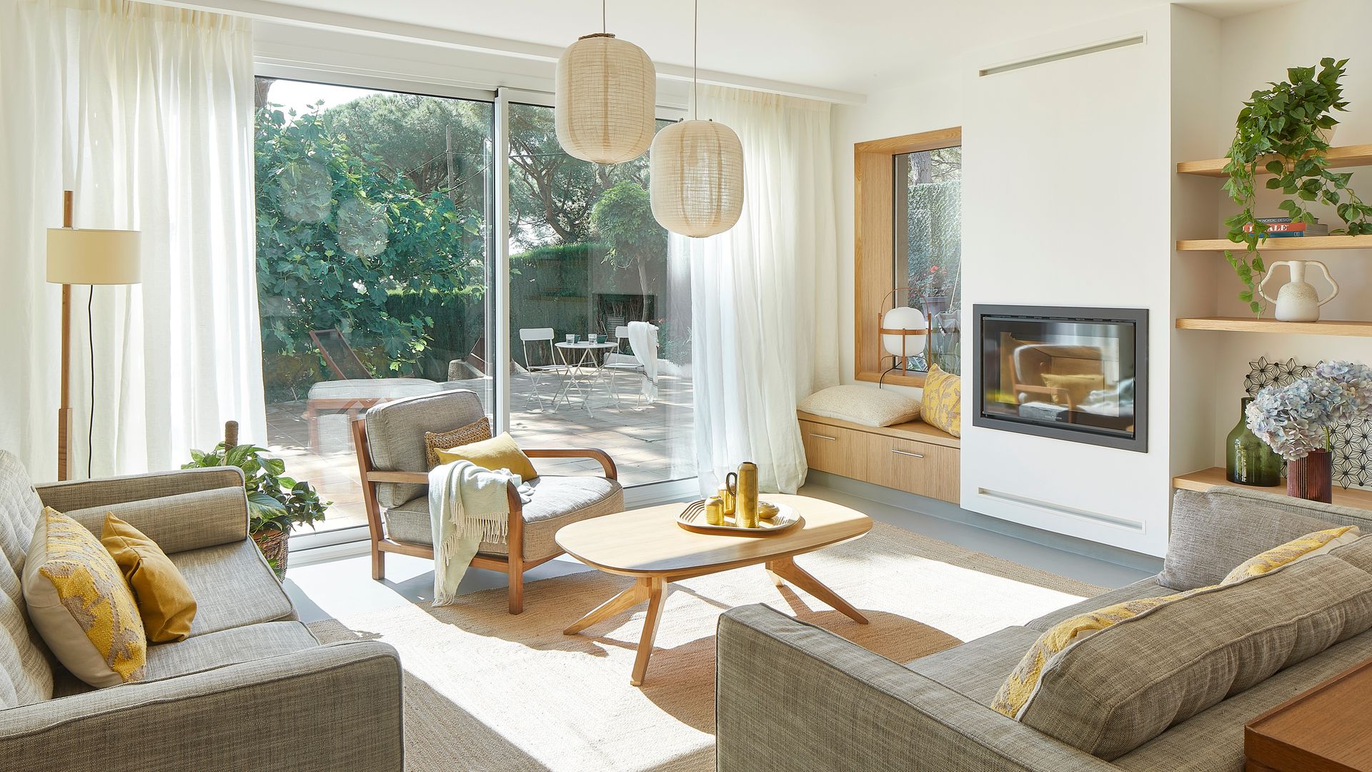 Salón con chimenea con salida al jardín lámparas de papel de techo, mesa de centro de madera, banco junto a la ventana con cojines y lámpara Cesta