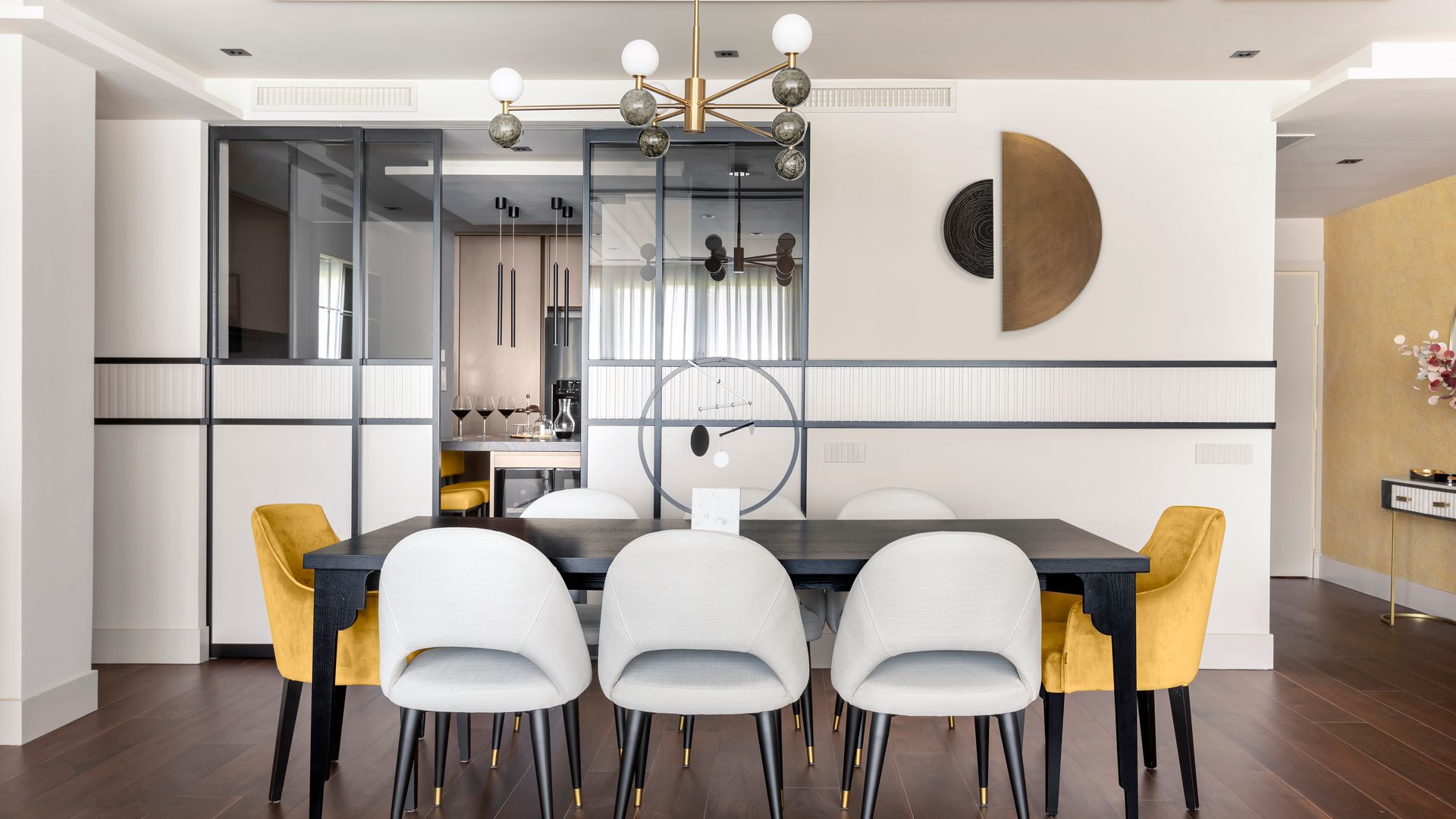 Comedor conectado con la cocina, suelo de madera oscuro, mesa de madera negra, sillas de comedor tapizadas en amarillo y blanco, lámpara de techo