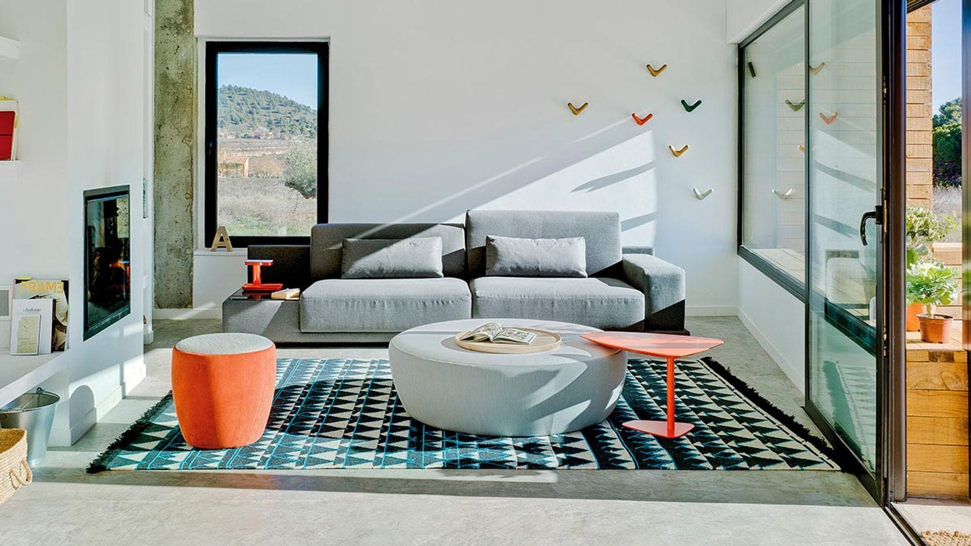 Salón con chimenea y salida al jardín, sofá gris, mesa de centro tapizada, alfombra y mesas auxiliares rojas