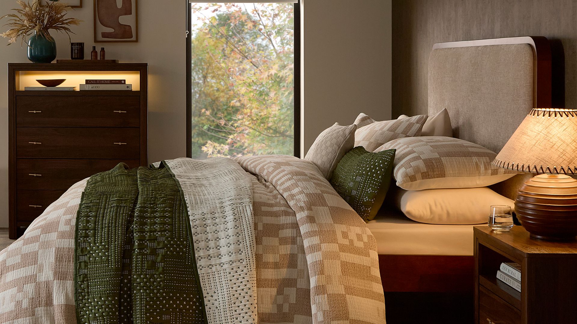 Dormitorio con manta y ropa de cama de invierno, cabecero tapizado, estor, cómoda, lámpara de sobremesa y en la cómoda encendidas