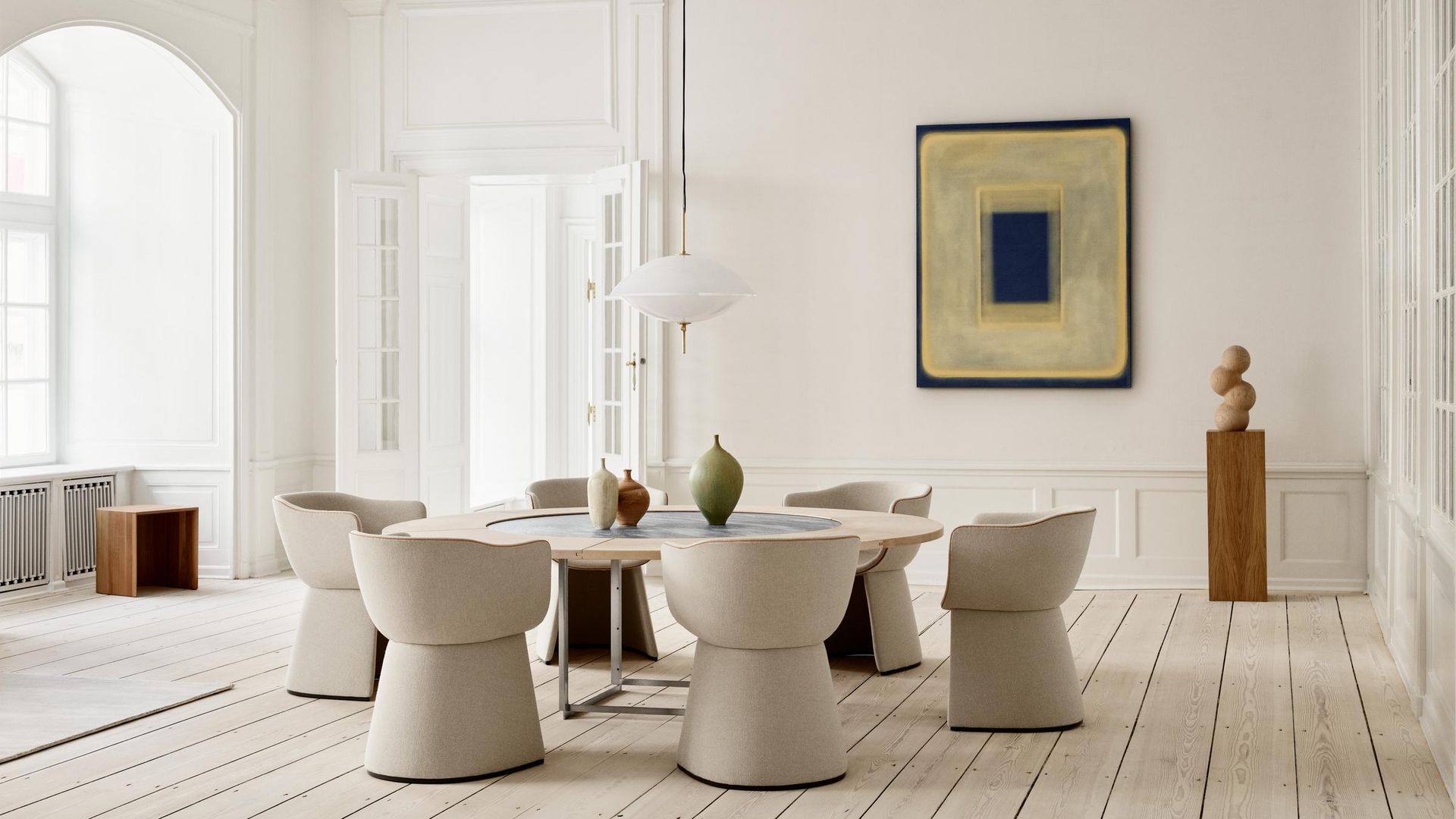 Comedor grande con mesa redonda XL, sillas tapizadas, lámpara de techo, suelo de madera, cuadro en la pared, puertas blancas con cuarterones de cristal