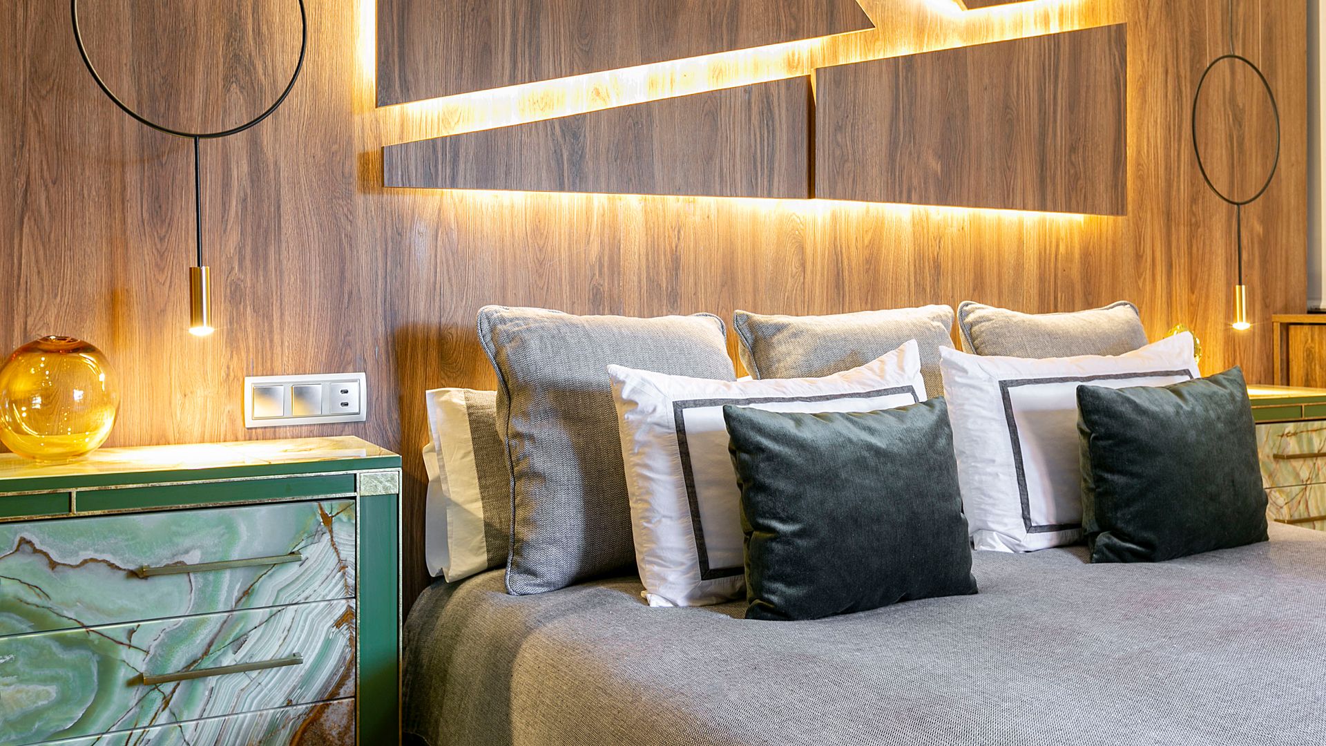 Dormitorio con pared de madera, retroiluminada, cama con cojines y cuadrantes, mesita de noche de color verde, lámparas de techo, ropa de cama gris