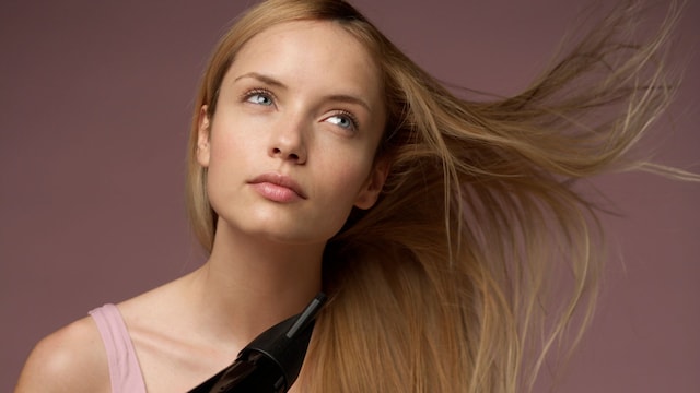 Mujer rubia con ropa interior rosa sentada en el suelo mirando al techo mientras se seca el cabello con un secador negro