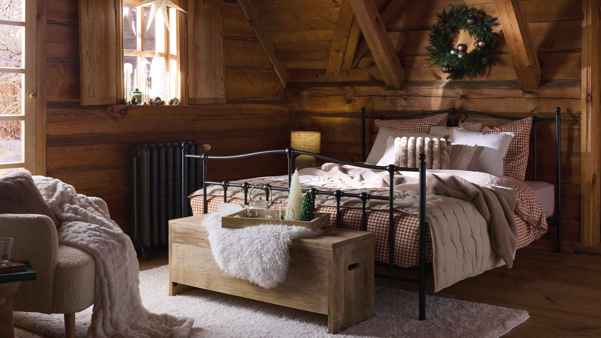 Dormitorio de cabaña con paredes y techo de madera, cama de hierro negro, corona de Navidad, estrella de navidad en la ventana, funda de cuadros rojos y blancos. alfombra de pelo largo, baúl de madera a los pies de la cama, butaca con manta