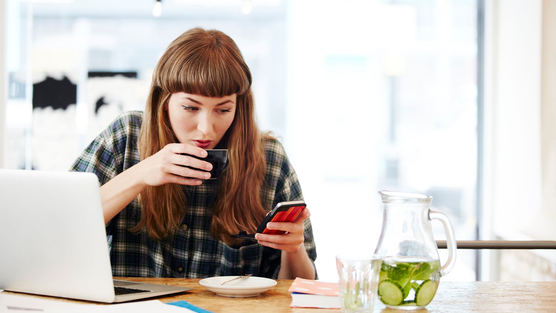 mujer tomando un café mientras mira su móvil y tiene abierto el portátil
